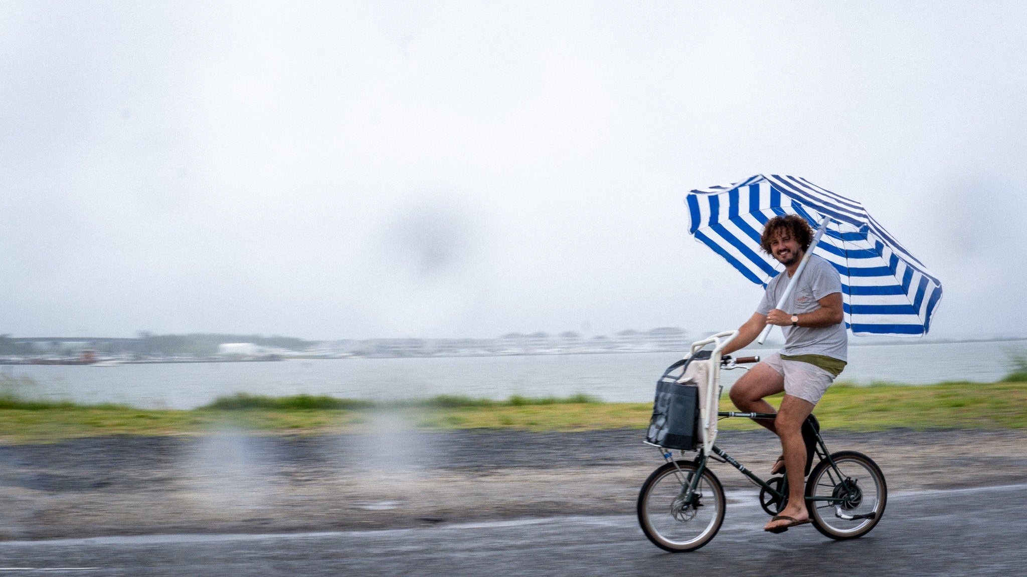 Person riding a bicycle with a striped umbrella on a rainy day by a lake.